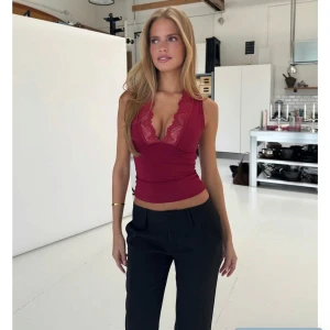 Vinröd ärmlös topp med spets - Topp från Venderbys, helt oanvänd med prislapp kvar. Vinröd ärmlös topp med djup v-ringning och breda spetsdetaljer längs urringningen. Figurnära passform och croppad längd, perfekt att styla med höga byxor. Toppens material är stretchigt och mjukt för en bekväm känsla.