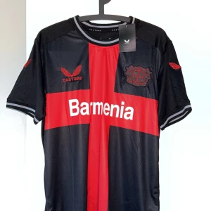 Bayer Leverkusen Wirtz matchtröja - Snygg Bayer Leverkusen fotbollströja från Castore med Wirtz och nummer 10 på ryggen. Svart bas med röd korsdesign och vita detaljer, klubbmärke och sponsortryck. Kortärmad modell i lätt och ventilerande material, perfekt för match eller träning.