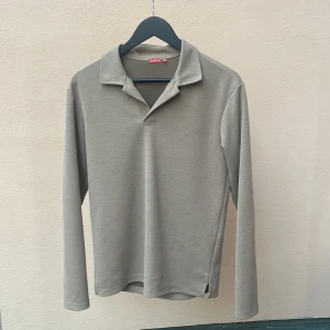 Beige långärmad pikétröja från RYLS - Säljer en stilren beige pikétröja från RYLS med klassisk krage och lång ärm. Tröjan har en enkel och clean look utan knappar framtill och är tillverkad i ett mjukt bomullsliknande material. Perfekt för dig som gillar minimalistisk stil.