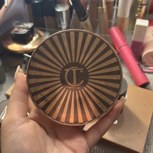 Charlotte Tilbury bronzer i guldig dosa - Snygg bronzer från Charlotte Tilbury i en lyxig rund guldig dosa med solstråle-design och spegel. Pudret har en varm, gyllenbrun ton som ger en naturlig solkysst look. Perfekt för contouring eller att värma upp ansiktet. Kvittot och förpackningen finns.