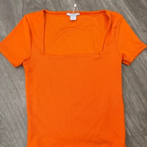 Orange topp med fyrkantig ringning - Snygg orange topp från Amiu med korta ärmar och en modern fyrkantig ringning. Perfekt att styla med jeans eller kjol för en färgstark look. Mjuk och stretchig känsla som sitter skönt på kroppen.