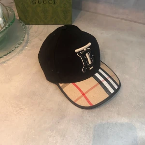 Burberry keps - Snygg svart keps från Burberry med klassiskt rutigt mönster på skärmen och justerbart band bak. Stor broderad logga framtill i vitt och svart. Perfekt accessoar för att lyfta din outfit med en lyxig touch.