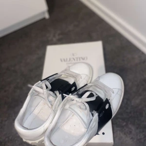 Valentino Garavani Open sneakers vit/svart - Snygga Valentino Garavani Open sneakers i vitt skinn med bred svart läderdetalj över vristen. Klassisk rund tå, vita snören och diskret logga på sidan. Perfekta för dig som vill ha en stilren och lyxig look med edge.