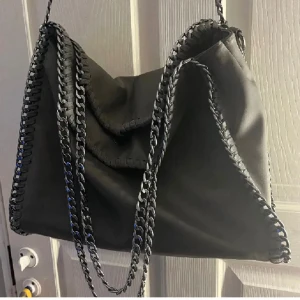 Grå axelväska med kedjeband Tiamo - Stilren grå axelväska med coola kedjor längs kanterna och som axelrem. Väskan har en mjuk, slouchy form och är tillverkad i skinnimitation. Perfekt för dig som gillar en edgy look och vill ha plats för allt du behöver.