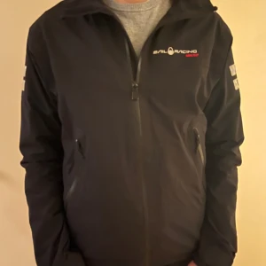 Svart vindjacka från Sail Racing - Svart vindjacka från Sail Racing med dragkedja framtill och två sidofickor med dragkedja. Jackan har logga på bröstet och detaljer på ärmarna. Perfekt för blåsiga dagar och snygg till jeans eller träningsbyxor.