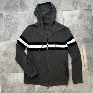 Grå hoodie från Massimo Dutti - Snygg grå hoodie från Massimo Dutti i extrafin bomull. Tröjan har dragkedja, huva med snörning och två ränder i vitt och mörkblått över bröstet. Perfekt för en chill och stilren look.