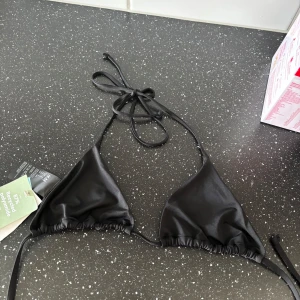 Svart trekantsbikini från H&M - Svart trekantsbikini från H&M med tunna knytband i nacken och ryggen. Klassisk och stilren modell i mjukt material, perfekt för stranden eller poolen. Tillverkad av 87% återvunnen polyamid och 13% elastan.