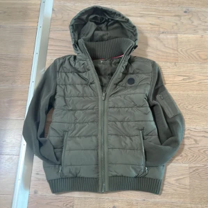 Grön cardigan  - Snygg olivgrön jacka med quiltad design och huva. Jackan har dragkedja framtill och ribbade muddar vid ärmslut och nederkant. Praktiska fickor med dragkedja och en extra ficka på ärmen. Perfekt för kyligare dagar!