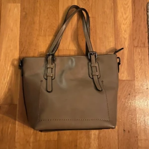 Beige axelväska med blommigt foder - Snygg beige axelväska med justerbara remmar och blommigt foder. Väskan har en dragkedja för säker förvaring och är perfekt för att bära dina dagliga nödvändigheter.