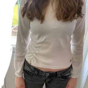 Vit ribbad topp från Marie Serneholt - Säljer en stilren vit ribbad topp från Marie Serneholt. Toppen är långärmad och har en snygg rynkdetalj på sidan. Tillverkad i ekologisk bomull med en liten del elastan för extra komfort. Perfekt för en avslappnad men chic look. Passar perfekt till low waist jeans som min modell har på sig på bilden. Tröjan är igentligen i storlek 146-152 men är stor i storlek. 