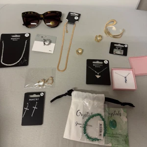 Smyckeskollektion med halsband, örhängen osv. - Värde: 600kr+. En samling av smycken inklusive ett silverfärgat halsband med triangelformad berlock, ett guldigt armband, och örhängen i både kors- och ringdesign. Inkluderar även ett grönt armband med kristaller och ett halsband med blå stenar. Osv. Både silvriga och guldiga smycken samt ett par solglasögon. Alla är oanvända, värde: 600kr+