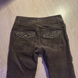 y2k Bruna low waist manchesterbyxor med pärldetaljer - Snygga bruna manchesterbyxor med pärldetaljer på bakfickorna. Byxorna har en slim passform och är högmidjade med dubbelknäppning fram. Perfekta för en stilren look.