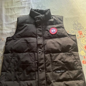 Svart dunväst från Canada Goose - Säljer en svart dunväst från Canada Goose med klassisk design och hög krage. Västen har knappar framtill och två stora fickor. Perfekt för kyliga dagar när du vill hålla värmen utan att kompromissa med stilen.