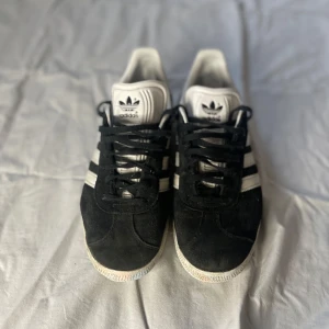 Svarta Adidas Gazelle sneakers - Säljer ett par klassiska svarta Adidas Gazelle sneakers med vita detaljer och snörning. Skorna har den ikoniska tre-randiga designen på sidorna och en vit sula. Perfekta för en stilren och sportig look.