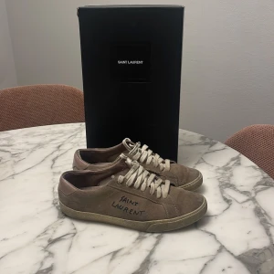 Beige mockasneakers från Saint Laurent - Snygga beige sneakers från Saint Laurent i mocka med snörning. Perfekta för en stilren look. Märkeslogga på sidan ger en exklusiv touch. Hör av er om ni har några frågor här dust bags.😊