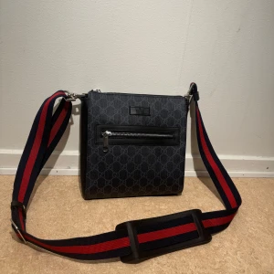 Svart axelväska med mönster - Snygg svart axelväska med ett diskret mönster och en justerbar axelrem i rött och blått. Väskan har en praktisk dragkedja framtill för enkel åtkomst till dina saker. Perfekt för att bära med sig det nödvändigaste med stil.