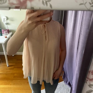Beige ärmlös blus med knyt - Säljer en luftig och elegant beige blus med knyt i halsen. Den är ärmlös och har en lätt plisserad design som ger en fin rörelse. Perfekt för varma dagar eller som ett lager under en kavaj.