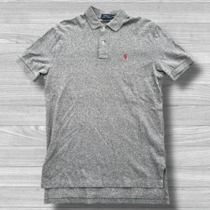 Ralph Lauren piké - Säljer en klassisk grå pikétröja från Ralph Lauren med den ikoniska röda logotypen broderad på bröstet. Tröjan har korta ärmar och en stilren krage med knappar. Perfekt för en avslappnad men stilfull look.