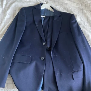 Mörkblå kostym från Dressmann (kostymbyxor ingår) - Säljer en stilren mörkblå kostym från Dressmann. Kavajen har en klassisk design med två knappar och fickor framtill. Passformen är slim fit, vilket ger en modern och elegant look. Perfekt för formella tillfällen. Enbart använd vid ett tillfälle.