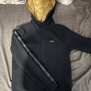 Svart boss kofta med guld detaljer varan sällsynt  - Snygg svart kofta från Boss med gulddetaljer och dragkedja. Jackan har en huva och är långärmad med ett stilrent märke på bröstet. Insidan är fodrad i guld och det finns dekorativa band längs ärmarna. Varan kostade över 2100kr i nypris nära 3000kr och säljs inte längre 