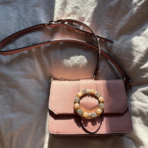 Rosa axelväska med dekorativt spänne - Snygg rosa axelväska med ett stort dekorativt spänne prytt med pärlor och stenar. Väskan har en justerbar axelrem och stängs med magnetknappar. Perfekt för att ge en touch av glamour till din outfit.