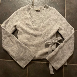 Grå omlottkofta från Gina Tricot - Säljer en mysig grå omlottkofta från Gina Tricot i premiumkvalitet. Koftan har långa ärmar och knytband i sidan för en snygg passform. Perfekt för kyliga dagar!