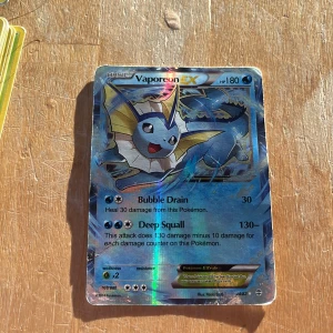 Vaporeon EX Pokémonkort - Säljer ett Vaporeon EX Pokémonkort med holografisk yta. Kortet har 180 HP och attacker som Bubble Drain och Deep Squall. Perfekt för samlare eller spelare som vill förstärka sin vattenbaserade lek.