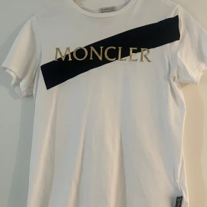 Moncler t-shirt - Vit Moncler t-shirt i Strl S med svarta och guldiga detaljer. Superfint skick då den endast är använd fåtal gånger. Köpt på room 1006 i Malmö men har tyvärr inte kvar kvittot. Nypris är ca 2800kr. 