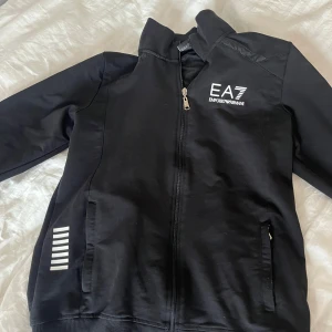 Svart tracksuit från Emporio Armani - Säljer en stilren svart jacka från Emporio Armani med dragkedja och vita detaljer. Jackan har långa ärmar och ribbade muddar. Perfekt för en sportig look. Om du vill köpa bara byxorna eller tröjan skriv så löser vi de.