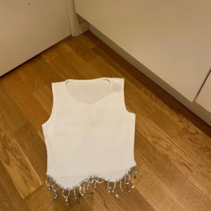 Vit ärmlös topp med pärldetaljer - Säljer en stilren vit ärmlös topp med dekorativa pärlor och fransar längs nederkanten. Perfekt för att ge en extra touch till din outfit. Passar bra till både jeans och kjol.
