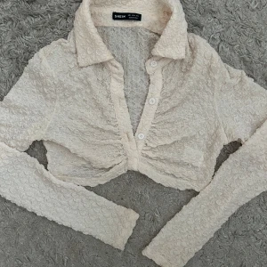 Beige croppad blus från Shein - Säljer en beige croppad blus från Shein med långärmad design och knappar framtill. Blusen har en unik textur och en stilren krage. Perfekt för att ge en trendig touch till din outfit. Helt oanvänd, och lite cream vit färg!😍