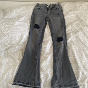 Grå bootcut jeans från Gina Tricot - Snygga grå bootcut jeans från Gina Tricot med slitna detaljer på knäna. De har en dragkedja och knapp framtill samt klassiska femficksdesign. Perfekta för en avslappnad stil.