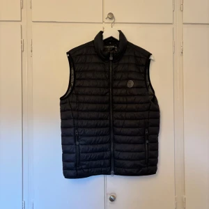 Svart quiltad väst från Marc O'Polo - Snygg svart quiltad väst från Marc O'Polo med dragkedja och hög krage. Perfekt för lager-på-lager under kyliga dagar. Västen har två praktiska fickor med dragkedja framtill.