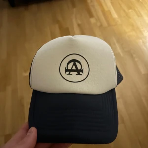 Vit och marinblå keps från All Cap One  - Snygg keps i vit och marinblå färg med en stilren logotyp framtill. Kepsen har ett nätmaterial baktill för bättre ventilation och en justerbar rem för perfekt passform. Kepsen är i 10/10 skick. Hör av dig 😉