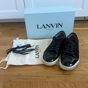 Lanvin Patent cap toe Navy skor 44 - Säljer ett par sneakers från Lanvin i storlek 44 då de blivit för små. De är i fint använt skick och väl omhändertagna. De har aldrig använts i regn/snö, det finns inga repor på den lackade tåkappan. Förutom själva skorna medföljer två dustbags, extra skosnören, skokartong och kvitto på köpet från NK i Stockholm. 