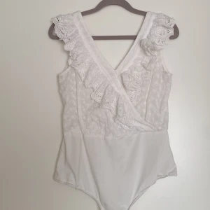 Vit spetsbody med volangdetaljer  - Säljer en elegant vit body med v-ringning och fina spetsdetaljer. Bodyn har volanger längs axlarna och omlott-design framtill.