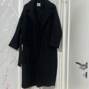 Svart kappa från Turkiet - Elegant svart kappa köpt i Turkiet med klassisk design. Den har långa ärmar och ett bälte i midjan för en stilren look. Perfekt för kyligare dagar.