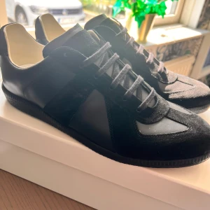 Svarta sneakers från Maison Margiela - Snygga svarta sneakers från Maison Margiela med en stilren design. Skorna har snörning och är tillverkade i en kombination av skinn och mocka. Perfekta för en trendig look. Aldrig använda! Hör av dig vid minsta fundering!