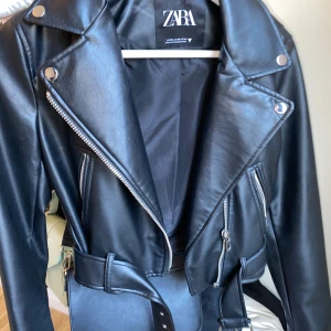 Svart skinnjacka från Zara - Snygg svart skinnjacka från Zara med dragkedja och bälte. Jackan har en klassisk bikerstil med långa ärmar och silverfärgade detaljer. Perfekt för att ge din outfit en edgy touch. Använd en gång förra sommaren. Inga defekter. Som ny.