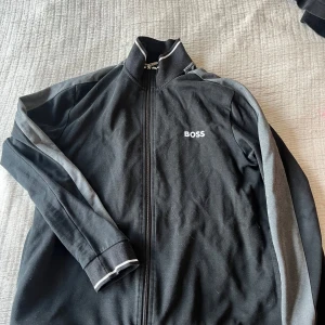 Svart fullzip från Boss - Säljer en stilren svart fullzip från Boss med grå detaljer längs ärmarna. Jackan har dragkedja framtill och en hög krage. Perfekt för en sportig look eller avslappnade dagar.