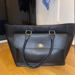 Svart axelväska med gulddetaljer - Snygg svart axelväska i syntetmaterial med guldfärgade detaljer. Väskan har en praktisk dragkedja och ett vridlås framtill. Insidan är rymlig med flera fack för enkel organisering. Får plats med dator o mycket annat. Perfekt för både vardag och speciella tillfällen. Några gropar på framsidan men inget som syns jättemycket i verkligheten. Säljer då ja inte använder den längre.(bilderna är från hon jag köpte av) skriv för mer bilder❤️❤️