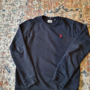 Mörkblå tröja från U.S. Polo Assn. - 🔵 Säljer en stilren marinblå tröja från U.S. Polo Assn med ett diskret rött broderi på bröstet. Tröjan har långa ärmar och rund halsringning, perfekt för en avslappnad look.  🔵 Priset är distribuerat vid snabb affär   🔵Ålder/storlek: 14-15 år