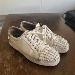 Louboutin vita med spikar fram och bak - Snygga vita sneakers från Louboutin med coola nitar på tån och hälen. Skorna har snörning och den ikoniska röda sulan. Perfekta för att ge din outfit en edgy touch. Slitna och utan sula, ligger en iläggs sula i nu. Köpta av Alex ceesay. Ta dem billigt. Storlek 46.