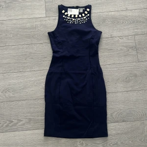 Marinblå klänning med pärldetaljer - Snygg marinblå klänning med pärldetaljer vid halsringningen. Klänningen är ärmlös och har en figurnära passform som framhäver silhuetten. Perfekt för en elegant kvällslook.