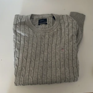 Grå stickad tröja från GANT - Säljer en stilren grå stickad tröja från GANT i stretchig bomull. Tröjan har ett klassiskt kabelstickat mönster och rund halsringning. Perfekt för en avslappnad men ändå snygg look. 🧶