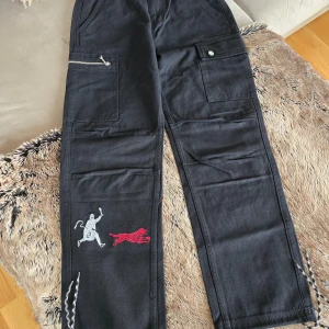 Svarta byxor med broderi från Galag - Snygga svarta byxor från Galag med coola broderier av en vit figur och en röd tiger. Byxorna har flera fickor och dragkedjedetaljer, samt snörning vid benen. Perfekta för en unik och stilren look. Helt nya.