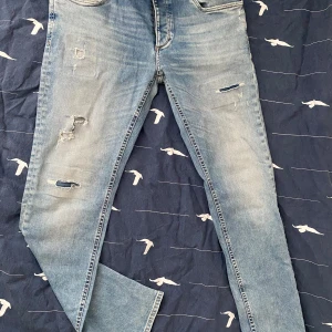 Blå slitna jeans - Säljer ett par blå jeans med slitningar och en klassisk femficksdesign. De har en dragkedja och knapp framtill. Perfekta för en avslappnad stil. Har inte använt dem så dem är helt fräscha.
