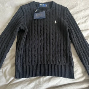 Svart stickad tröja från Ralph Lauren - Säljer en stilren svart stickad tröja från Ralph Lauren med kabelstickat mönster. Tröjan har långa ärmar och en rund halsringning. Perfekt för kyligare dagar och ger en klassisk look.