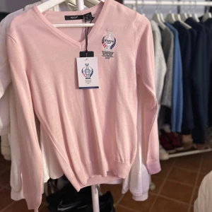 Rosa tröja från Abacus - Säljer en ljusrosa tröja från Abacus med Solheim Cup 2019 broderi på bröstet. Tröjan har en v-ringad design och långa ärmar, perfekt för golfentusiaster. Tillverkad i ett mjukt material för komfort.