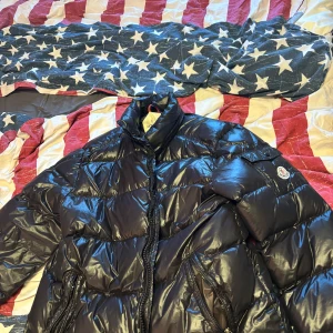 Svart pufferjacka från Moncler - Säljer en snygg svart pufferjacka från Moncler. Jackan har en glansig yta med dragkedja och en broderad logotyp på ärmen. Perfekt för kyliga dagar!